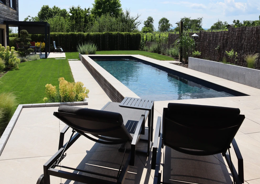 Jardin contemporain avec piscine rectangulaire à débordement, margelles en pierre claire, deux transats noirs design sur terrasse dallée